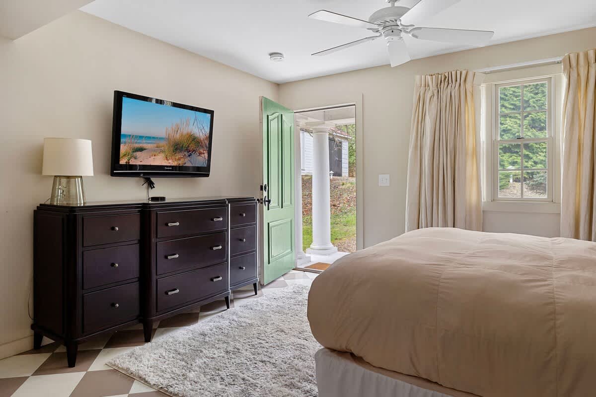 A cozy bedroom featuring a bed with light-colored bedding, a dark wood dresser with a wall-mounted flat-screen TV, and a soft area rug over a checkered floor. A light green door leads to an outdoor space, and a window provides natural light.