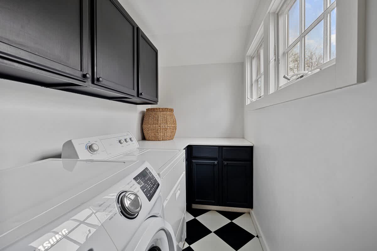 A bright and functional laundry room with crisp white walls, a striking black and white checkered floor, and modern white washer and dryer units. Black upper and lower cabinets provide ample storage, illuminated by a large window.
