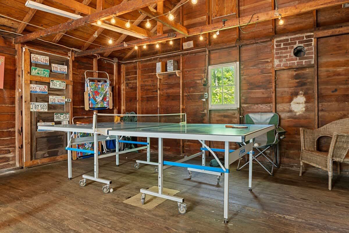 A closer view of the rustic game room with exposed wooden walls and ceiling, featuring a ping-pong table with blue accents. String lights illuminate the space, and various wall decorations, including license plates, add character.