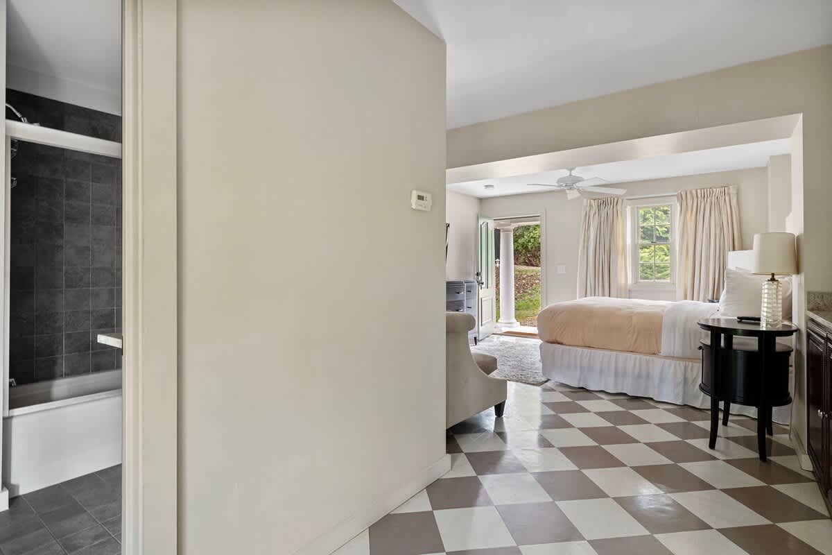 A transitional space with gray and white checkered flooring, offering a view into a bathroom with a shower on the left and a bedroom with a bed and ceiling fan on the right. The walls are painted a neutral beige.
