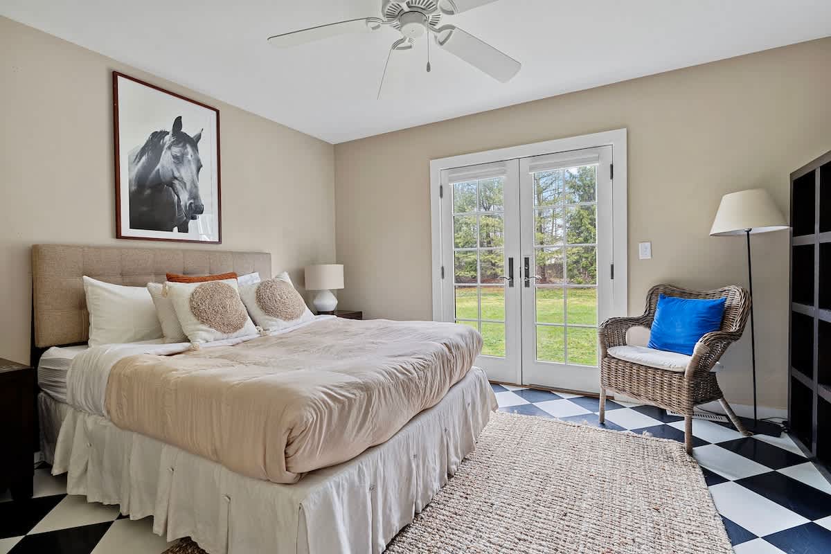 A bedroom featuring a queen-sized bed with a beige upholstered headboard and neutral bedding, a wicker chair with a blue pillow, and French doors opening to a grassy outdoor area. The room has a black and white checkered floor partially covered by a rug.