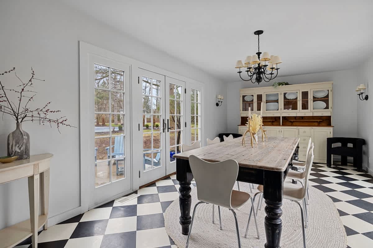 A spacious dining room featuring a large wooden table with black legs, surrounded by white chairs, set on a round jute rug over a black and white checkered floor. French doors open to an outdoor area, and a white hutch with glass doors is visible in the background.