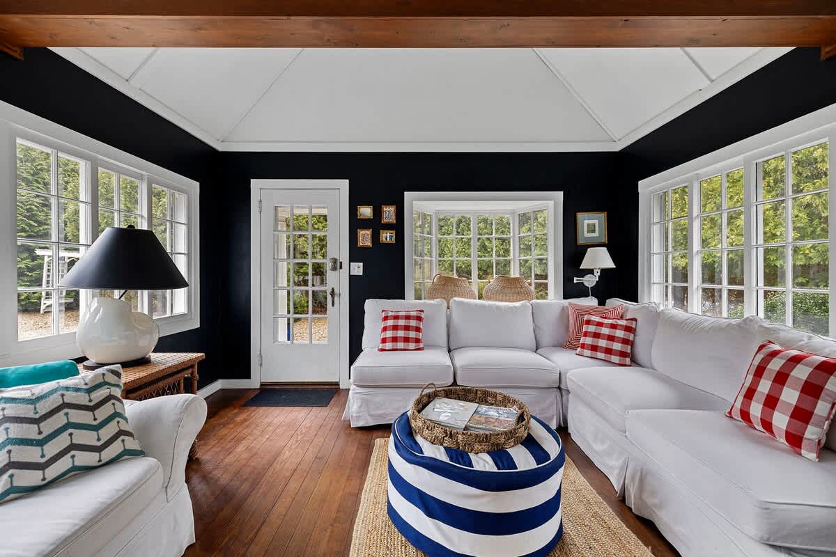 Another perspective of the sunroom shows white slipcovered sectional sofas with red and white checkered pillows, dark blue walls, and a vaulted white ceiling with wooden beams. A blue and white striped ottoman serves as a coffee table on a natural fiber rug.