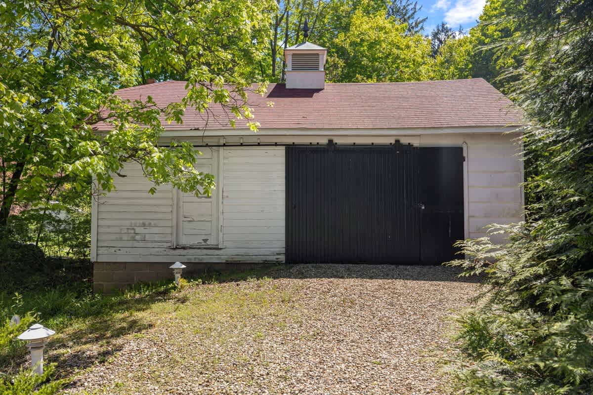 A rustic white barn-style garage with a red roof and a cupola, featuring large black sliding doors. The building is surrounded by green trees and a gravel driveway.