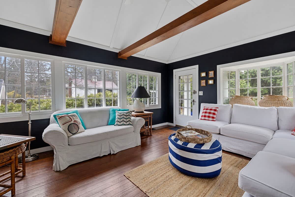 A bright sunroom features dark blue walls, white vaulted ceilings with exposed wooden beams, and multiple large windows overlooking greenery. White slipcovered sofas are adorned with colorful throw pillows, and a striped ottoman sits on a natural fiber rug.