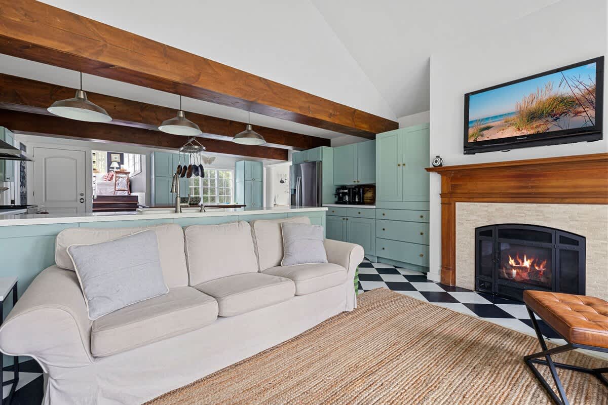 An open-concept living area with a light-colored sofa facing a fireplace with a wooden mantel and a mounted TV. The space opens into a kitchen with light blue cabinets, a breakfast bar, and exposed wooden beams.