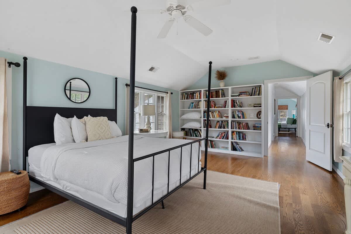 A spacious bedroom featuring a black four-poster bed with white bedding, light blue walls, and warm hardwood floors. A built-in white bookshelf filled with books and a round mirror above the bed add character.