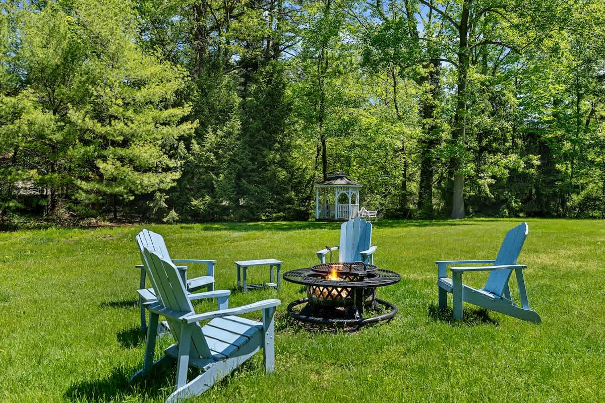 Light blue Adirondack chairs are arranged around a decorative black metal fire pit on a well-maintained green lawn. Tall, leafy trees provide a natural backdrop under a bright sky.