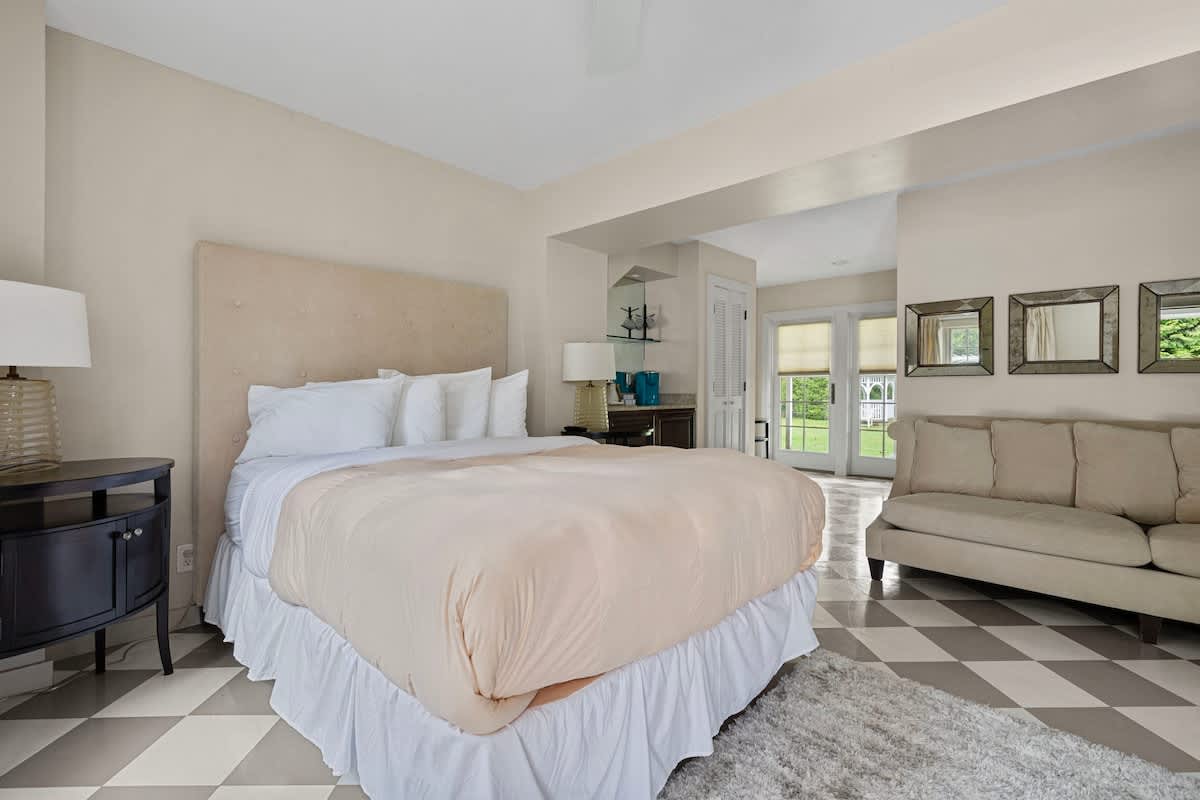 A spacious bedroom with a large bed featuring a beige upholstered headboard and white bedding, set on a distinctive gray and white checkered floor. A light-colored sofa and French doors leading to an outdoor area are also visible.
