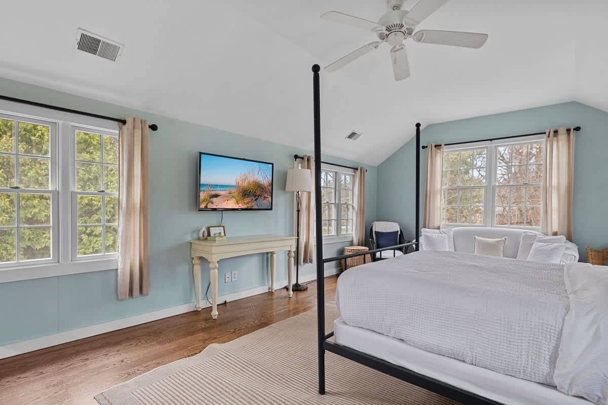 This bright bedroom showcases a black four-poster bed with white bedding, light blue walls, and hardwood floors. Multiple windows with light-colored curtains and a wall-mounted TV create a comfortable space.