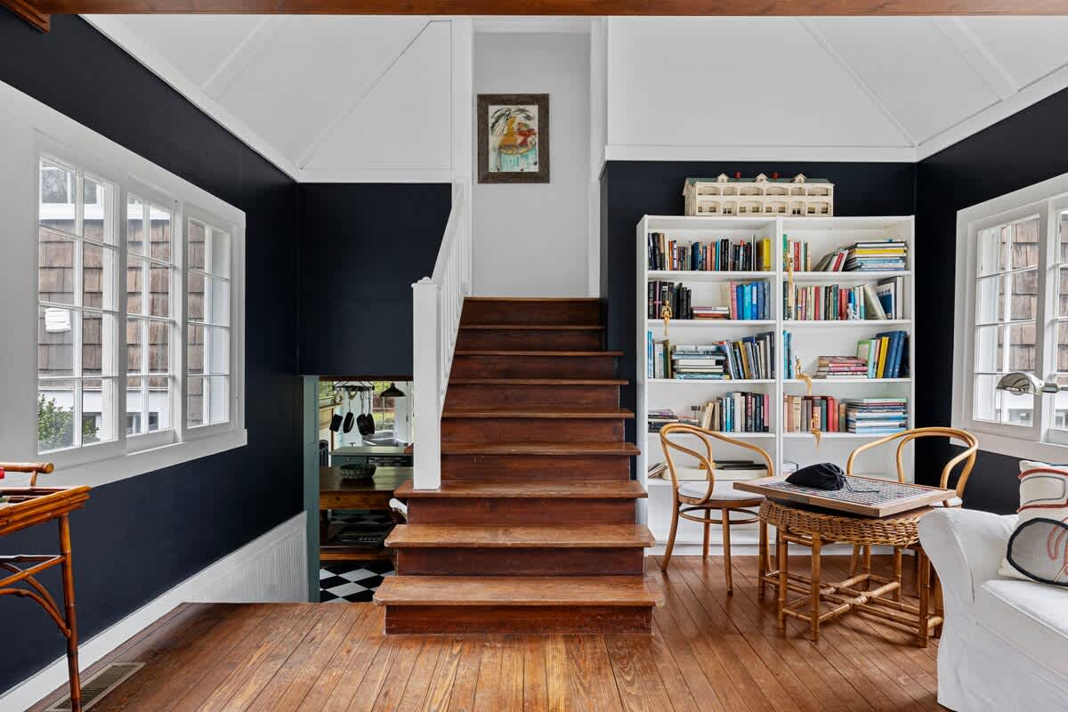 A wooden staircase with white railings ascends from a dark-walled living space, featuring a large white bookshelf filled with books. Hardwood floors and multiple windows are visible, with a small table and chairs in front of the bookshelf.