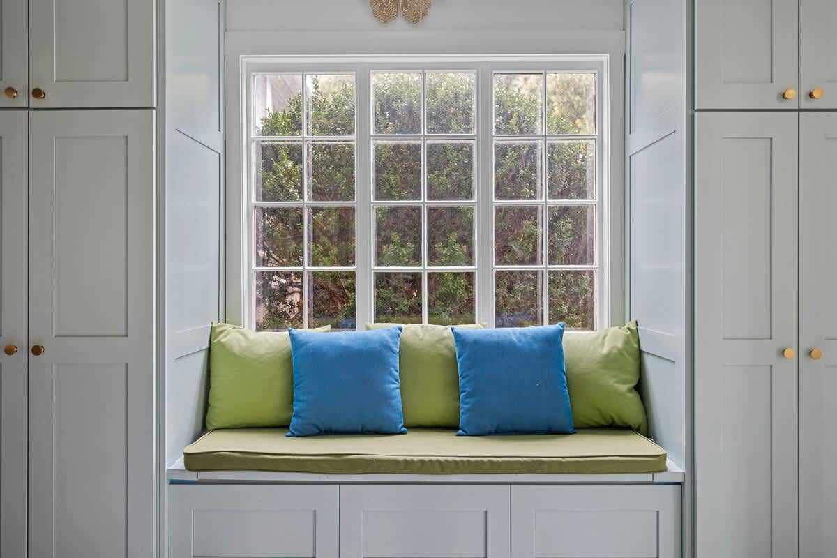 A cozy window seat with light blue built-in cabinetry, a green cushion, and blue throw pillows, flanked by matching tall cabinets. The large grid-patterned window offers a view of lush greenery outside.