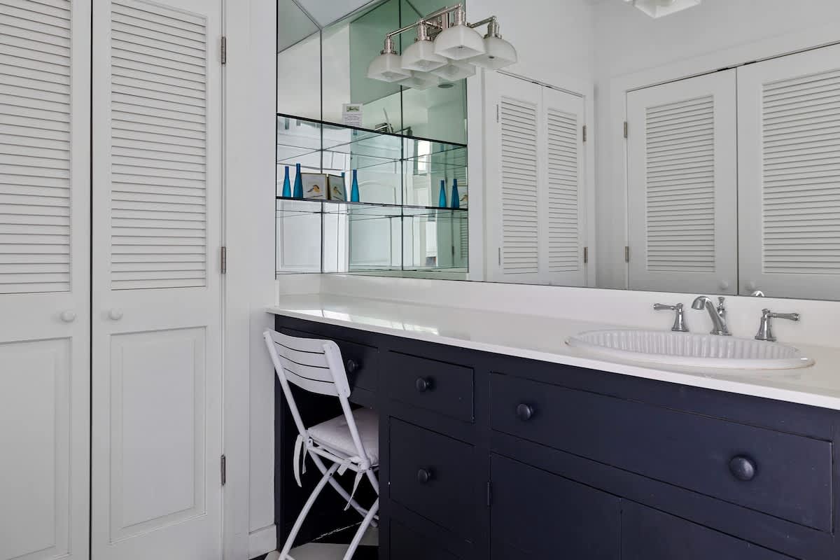 This bathroom features a dark blue vanity with a white countertop and an integrated sink, complemented by a large mirrored wall and white louvered closet doors. A white folding chair is neatly tucked under the vanity.
