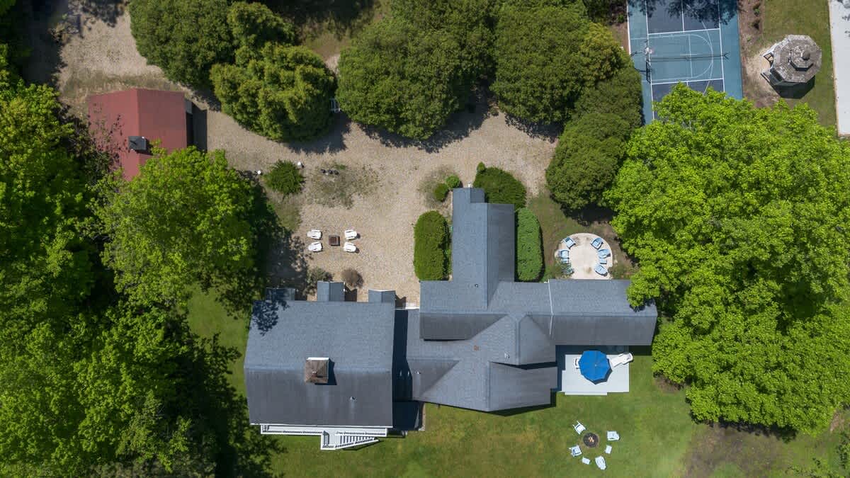 Overhead aerial view showcasing the property's layout, including the main house with a dark roof, surrounding green lawns, and a glimpse of the tennis court and other outdoor seating areas.