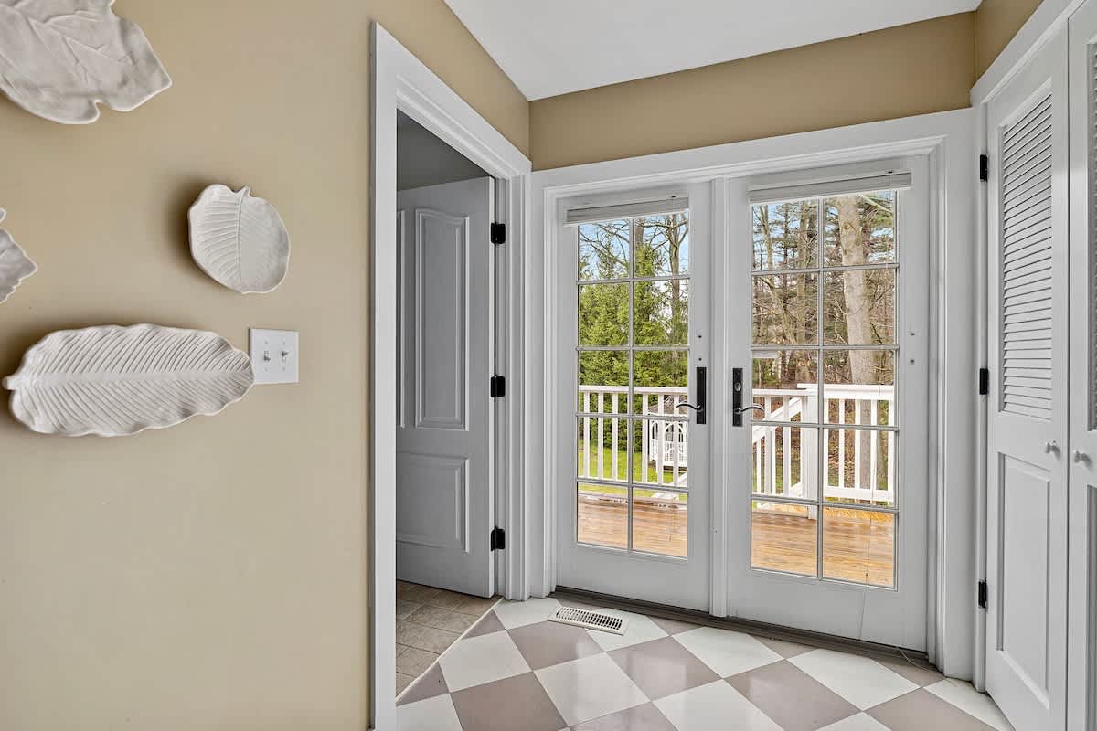 An interior view of a transition area with gray and white checkered flooring, featuring white French doors that open onto an outdoor deck or balcony. Decorative white leaf-shaped wall art adorns the neutral-colored wall.