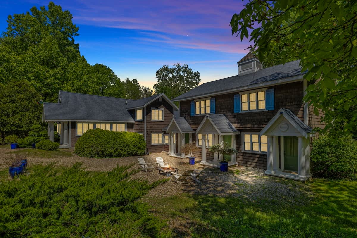 Charming shingle-sided house illuminated at dusk, featuring multiple gabled entrances with white columns and blue shutters, with a gravel fire pit area in the foreground.