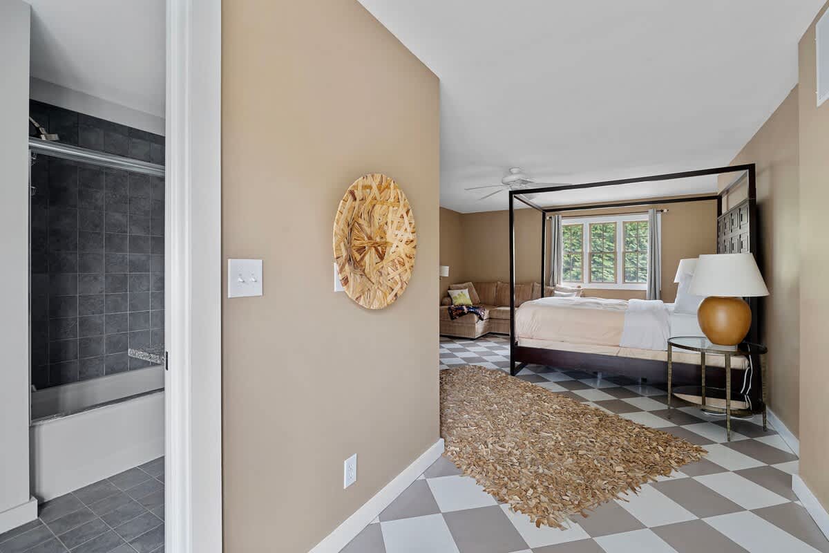 A hallway with light tan walls and a checkered floor, leading into a bedroom with a four-poster bed. A bathroom entrance with a shower/tub combo is visible on the left, and a decorative woven wall hanging adorns the wall.