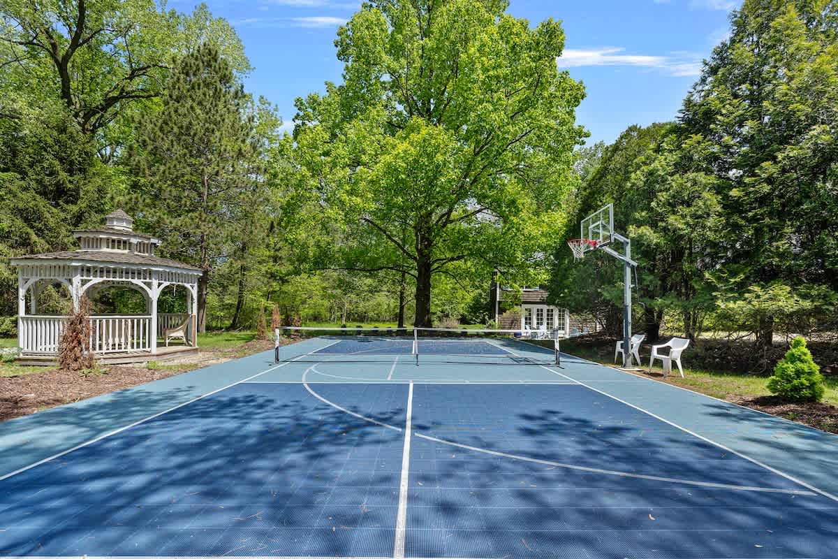 A vibrant blue and green multi-sport court, marked for pickleball or tennis, features a net and a basketball hoop. A white gazebo is visible in the lush green background, surrounded by mature trees.