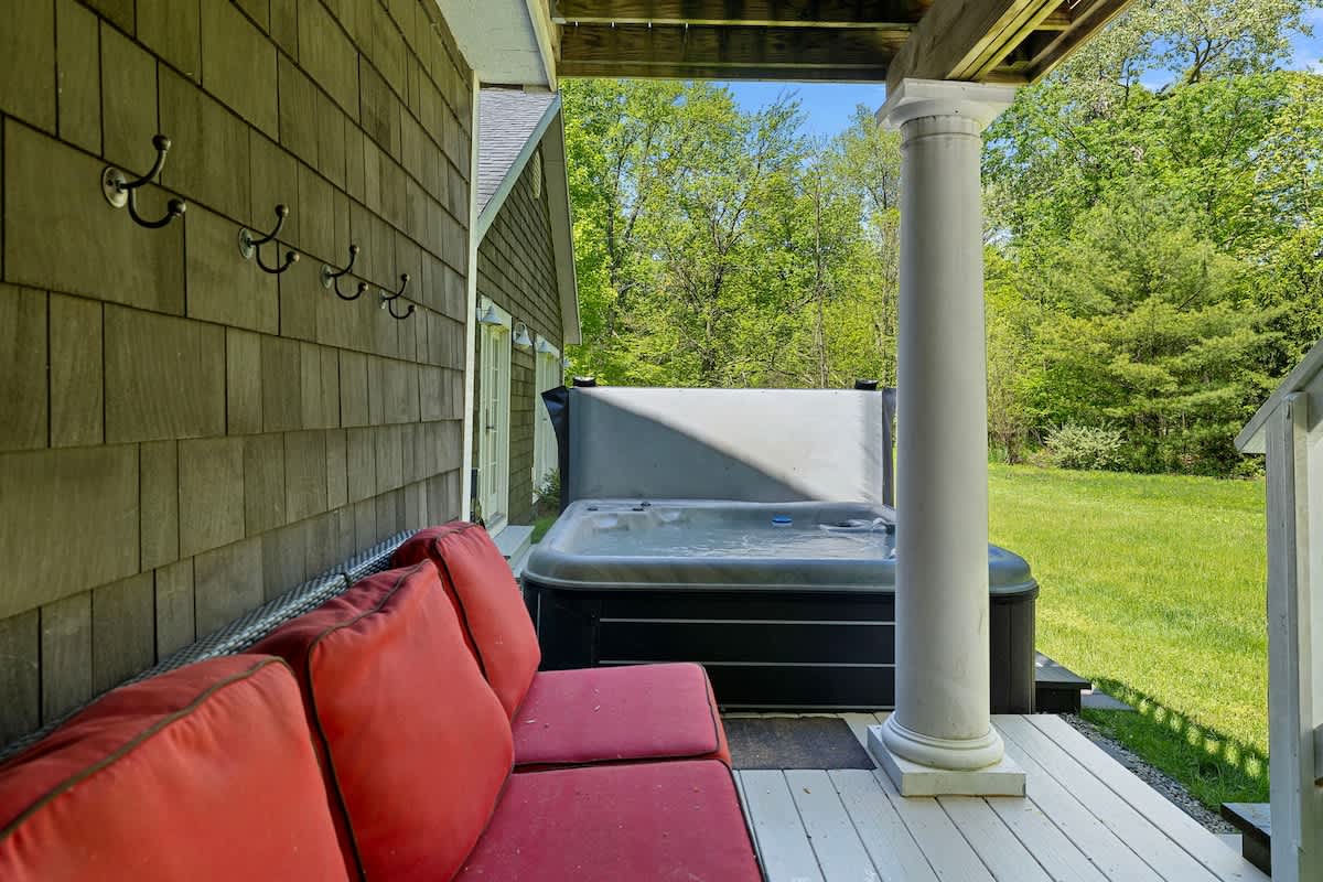 A covered outdoor deck with gray flooring, featuring a hot tub with a cover and a dark wicker sofa with red cushions. White columns support the overhead structure, and a lush green yard is visible.