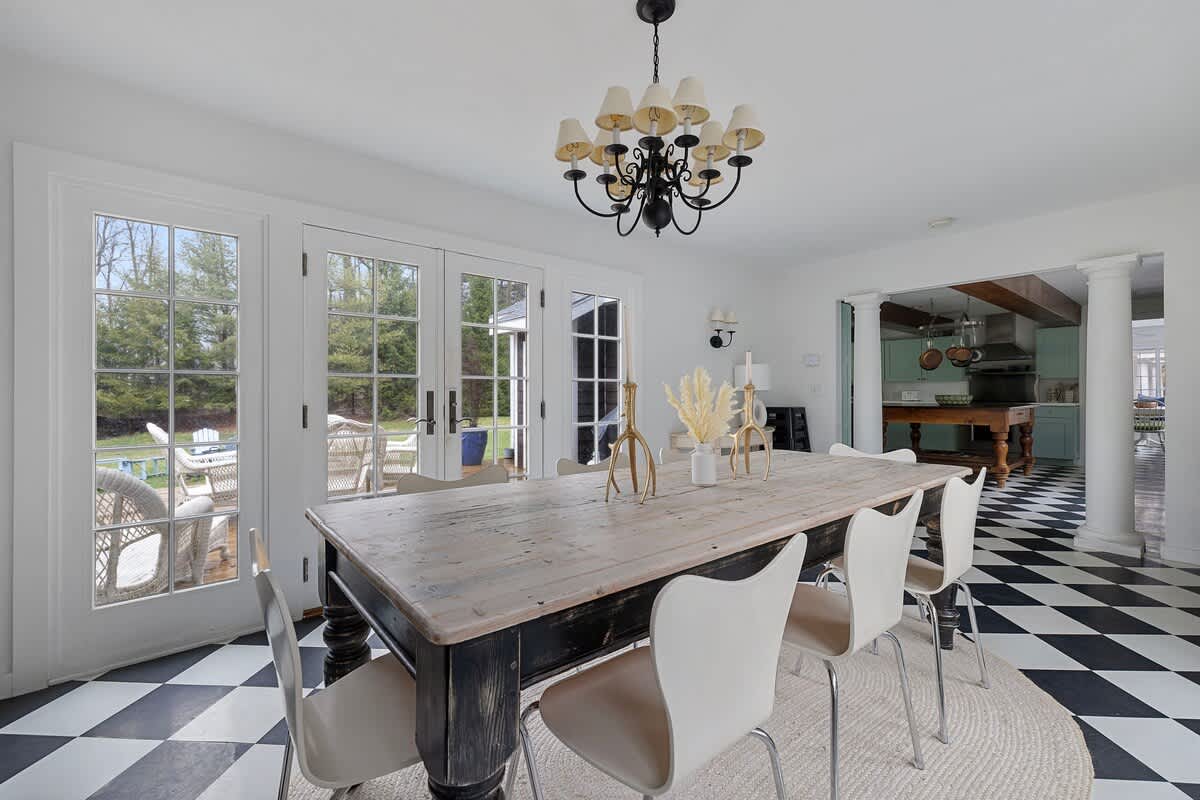 This dining room features a large wooden table with white chairs on a jute rug over a black and white checkered floor. French doors lead outside, and an open archway reveals a kitchen with light blue cabinets and exposed wooden beams.