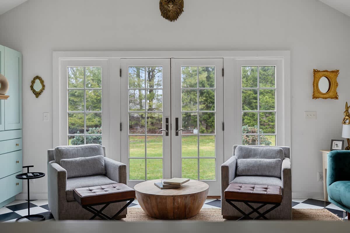 A bright sitting area features two gray armchairs with leather ottomans facing French doors that open to a grassy yard. A round wooden coffee table sits between the chairs on a natural fiber rug, with light green cabinets visible in the background.