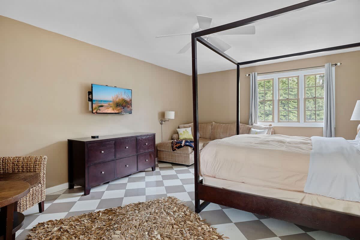 A spacious bedroom with light tan walls and a checkered floor, featuring a dark wood four-poster bed with light bedding, a wall-mounted TV, and a dresser. A small seating area with a sofa and a window is visible in the background.