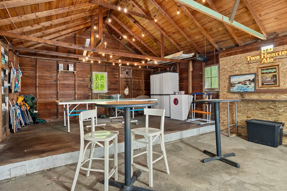 A spacious game room with exposed wooden beams and walls, featuring string lights and a concrete floor. A ping-pong table is central, with high-top tables and chairs, and a refrigerator/freezer unit in the background.