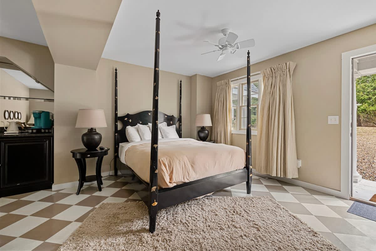 This bedroom showcases a black four-poster bed with gold detailing, light beige walls, and a checkered floor. A window with light curtains and an exterior door provide access and natural light.