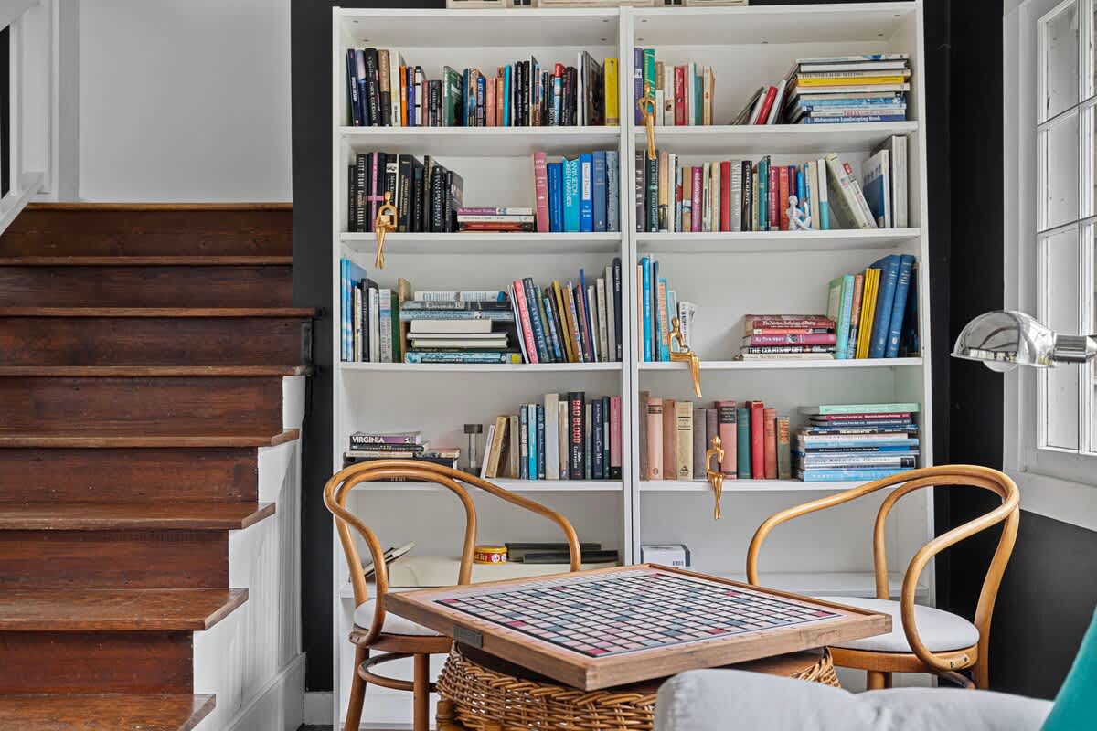 A detailed view of the white built-in bookshelf, fully stocked with books and decorative items, stands beside a wooden staircase. Two bentwood chairs and a wicker table with a board game are placed in front, on hardwood floors.