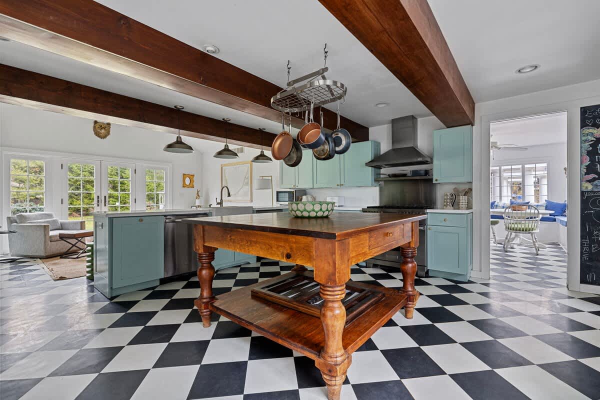 An expansive kitchen features light green lower cabinets, a large wooden island, and a stainless steel range with a hood. The black and white checkered floor contrasts with the white walls and exposed wooden beams, with French doors leading to an outdoor area and a glimpse of another room.