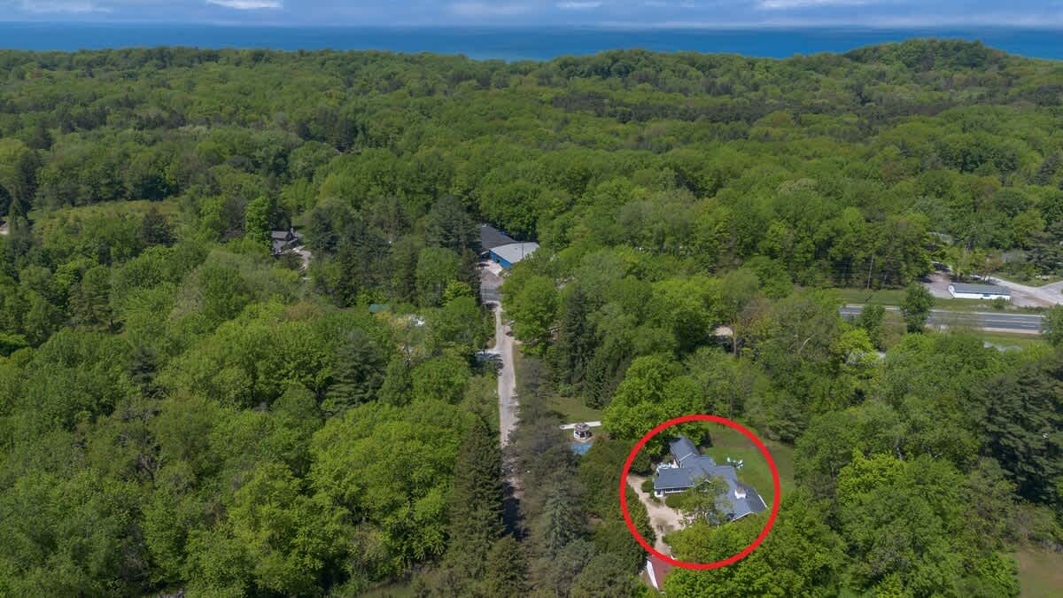 An aerial view of a property nestled within a dense green forest, with the main house highlighted by a red circle, a winding road, and a large body of water visible on the distant horizon.