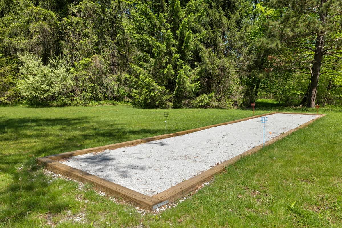 A well-maintained bocce ball court, featuring a light gravel surface framed by wooden timbers, is nestled within a vibrant green lawn, bordered by dense evergreen trees.