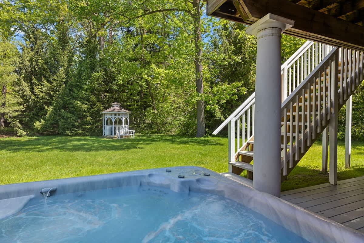 A bubbling hot tub is positioned on a wooden deck, with white stairs leading up to a house. In the background, a white gazebo sits on a lush green lawn surrounded by tall trees.