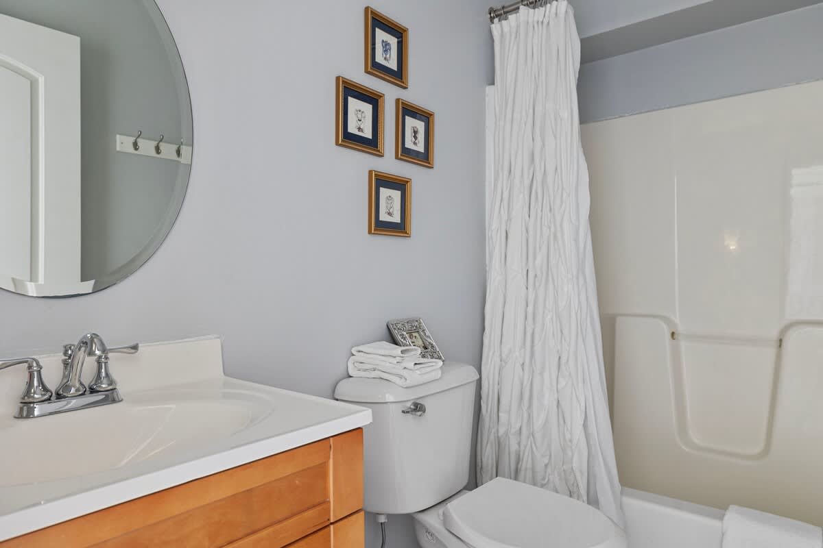 A light-colored bathroom with a single wooden vanity, a round mirror, a white toilet, and a shower/tub combo with a white ruffled shower curtain. The walls are painted a soft blue-grey.
