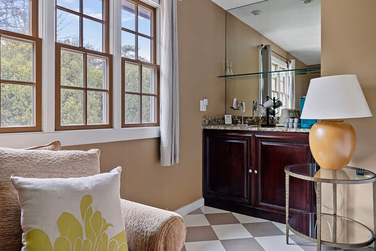 A cozy corner of a room with light tan walls and a checkered floor, featuring a wet bar with dark wood cabinetry, a granite countertop, and a mirror backsplash. A large window with multiple panes overlooks lush greenery.