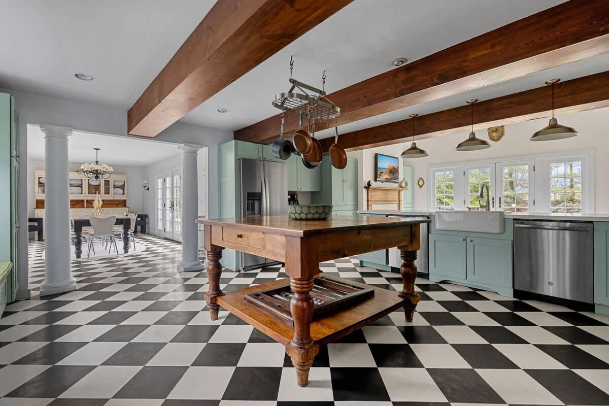 A wide view of the kitchen showcases the black and white checkered floor, light green cabinets, and a large wooden island. Exposed wooden beams span the ceiling, and an opening with white columns leads to a dining area with a chandelier.