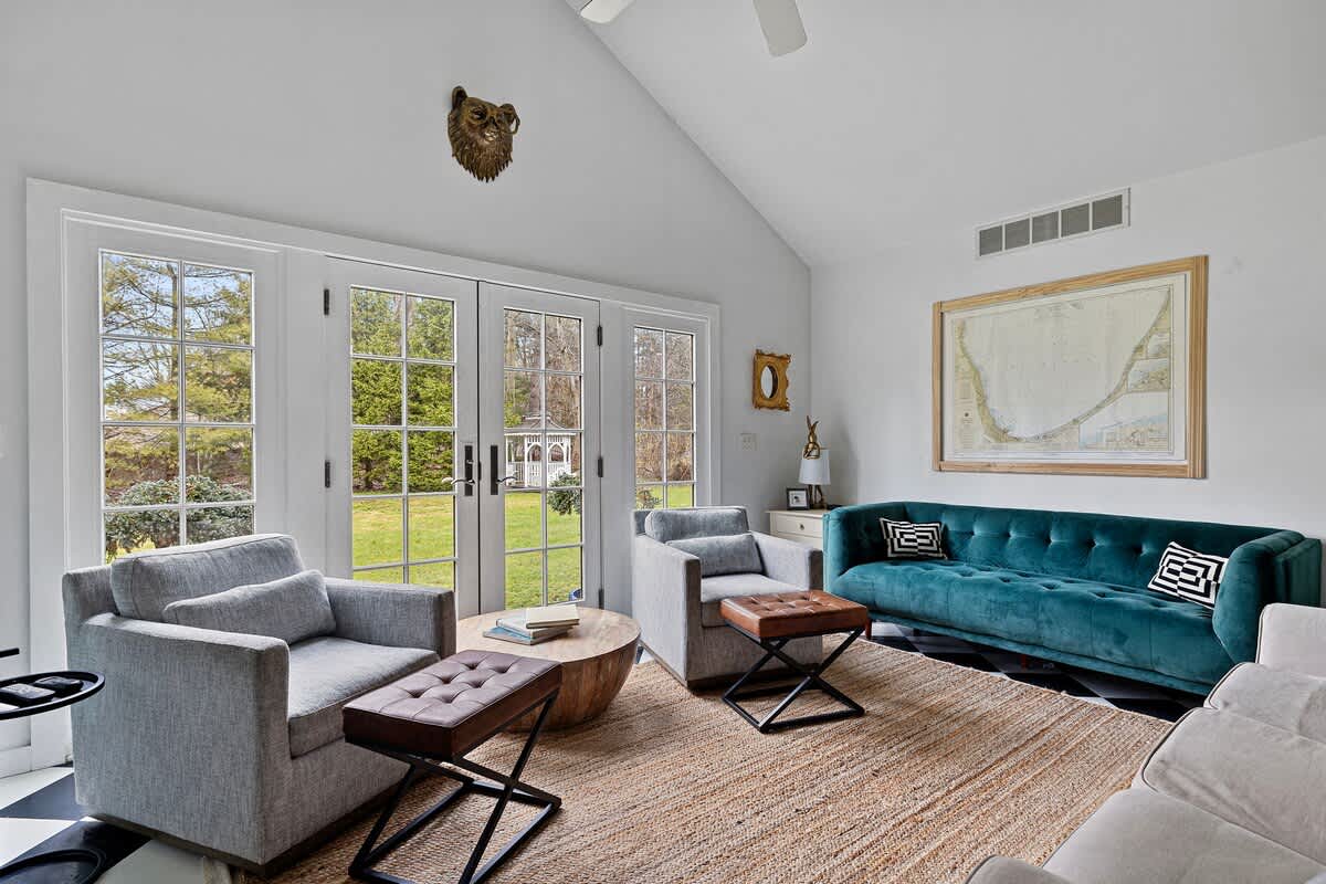 A bright living room with a vaulted ceiling, featuring a teal velvet sofa, two grey armchairs with ottomans, and a round wooden coffee table, all arranged on a large jute rug. French doors provide a view of the outdoors.