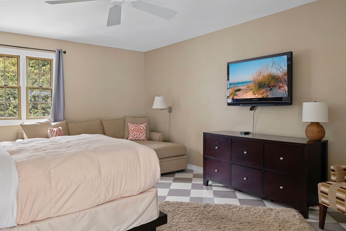 A bright bedroom with a bed featuring light bedding, a dark wood dresser with a wall-mounted TV, and a chaise lounge. The room has a checkered floor, a ceiling fan, and a window with light-colored curtains.