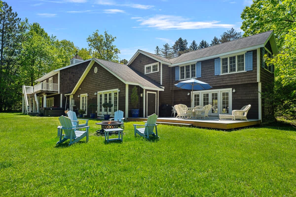 The back of a shingle-sided house features a wooden deck with outdoor dining furniture and an umbrella. On the expansive green lawn, light blue Adirondack chairs surround a black fire pit, with lush trees in the background.