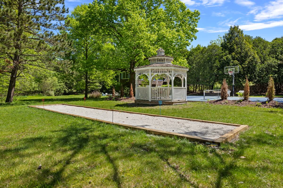 A long, rectangular bocce ball court with a sandy surface and wooden borders is set on a manicured green lawn. A white gazebo and a blue sports court are visible in the background, framed by trees.