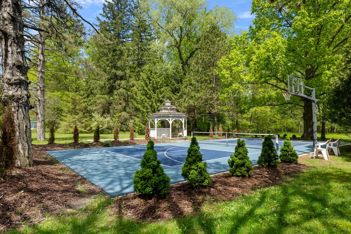 Expansive outdoor recreation area featuring a blue multi-sport court for tennis or pickleball, a white gazebo, and a basketball hoop, surrounded by lush trees and landscaping.