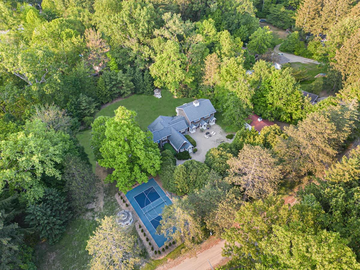 Aerial view of a large house with a dark roof nestled among lush green trees, featuring a blue outdoor tennis or pickleball court and a gravel driveway.