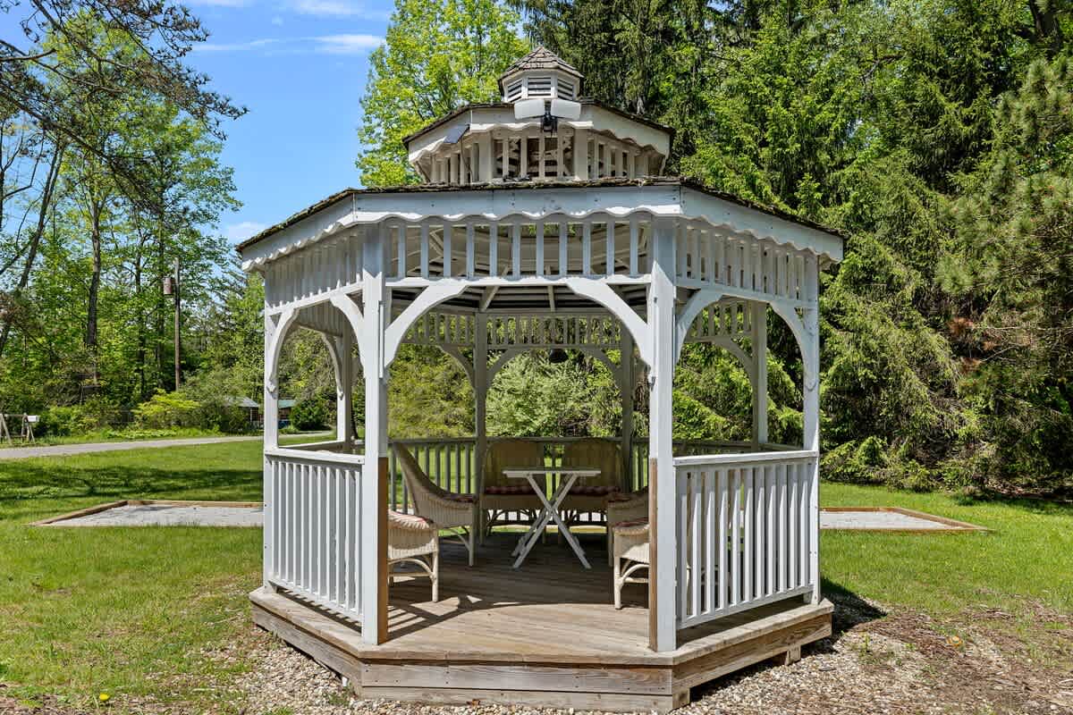 A charming white wooden gazebo with intricate lattice work and a multi-tiered roof stands on a wooden deck, furnished with wicker chairs and a small table, set amidst a green lawn.
