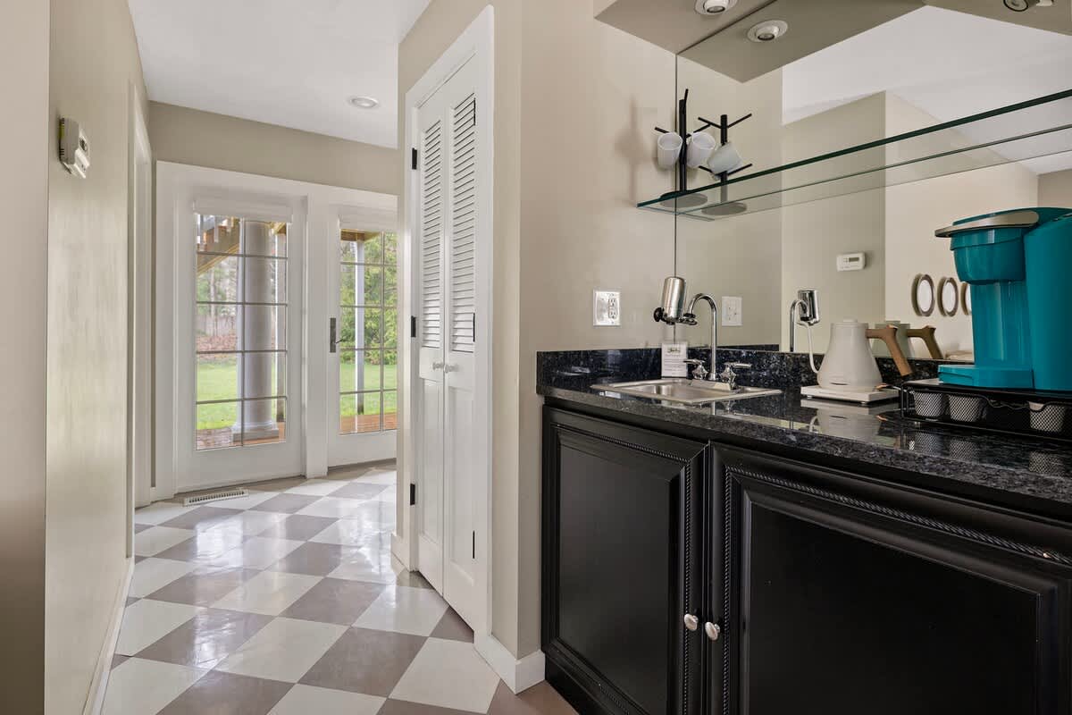 A compact kitchenette or wet bar area featuring sleek black cabinets, a dark granite countertop, a small sink, and a mirrored backsplash. A coffee maker and kettle are conveniently placed on the counter.