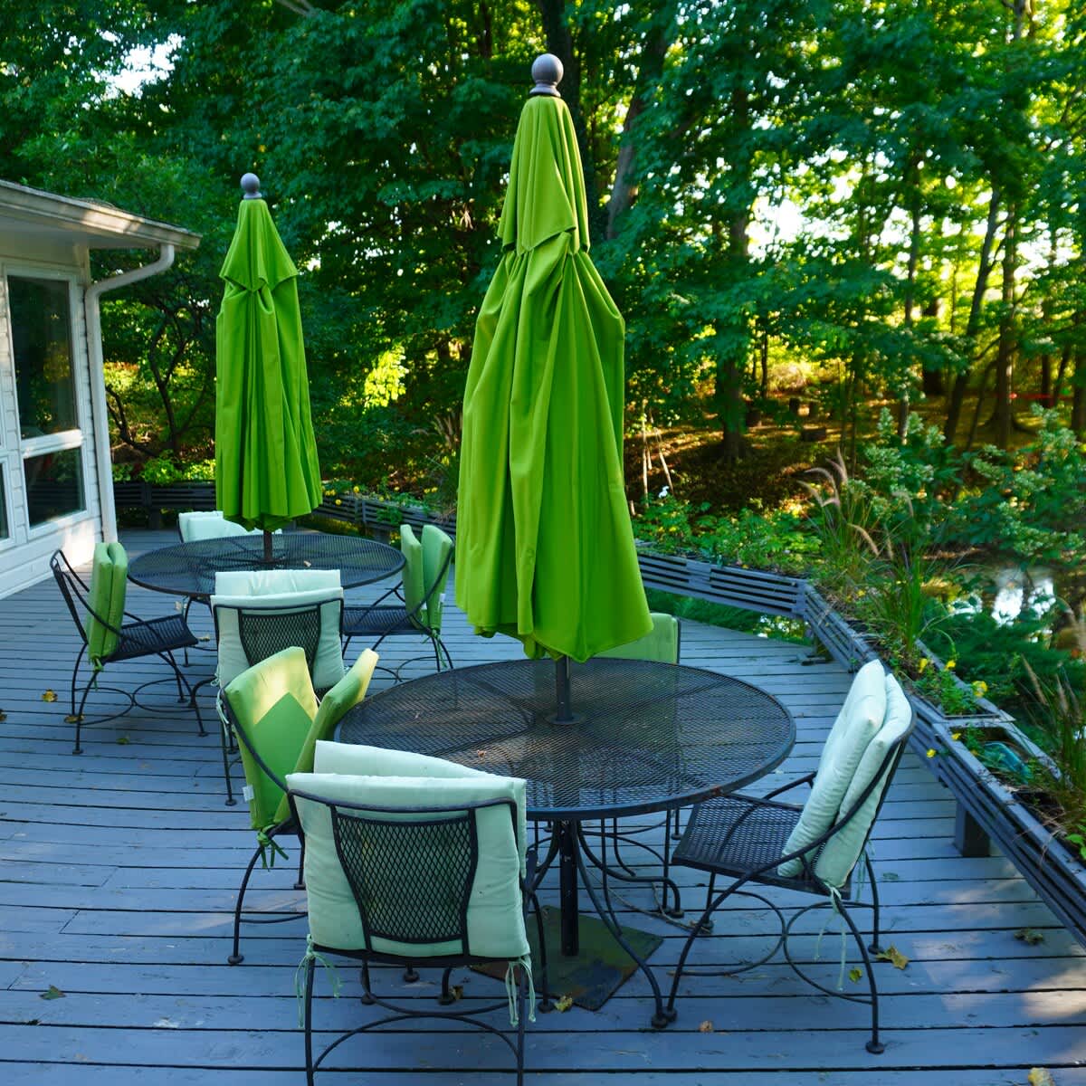 Spacious outdoor deck featuring multiple black metal tables and chairs with light green cushions and large matching umbrellas. The deck is surrounded by lush green trees and overlooks a serene body of water.