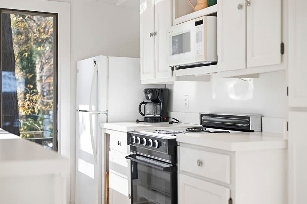 Bright, compact kitchen featuring white shaker cabinets, white countertops, and a white refrigerator. Appliances include a black oven/stove, a microwave, a coffee maker, and a toaster oven, all well-lit by natural light.