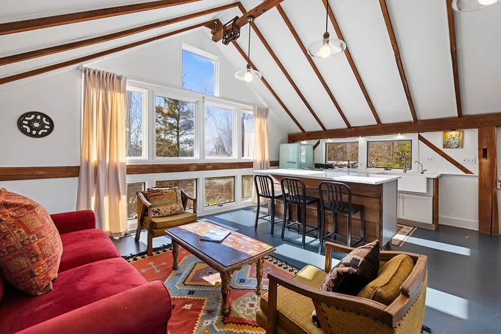 Open-concept living area and kitchen with a high vaulted ceiling featuring exposed wooden beams. The living space has a red sofa, two armchairs, and a colorful rug, while the kitchen features a central island with bar stools and large windows overlooking the outdoors.