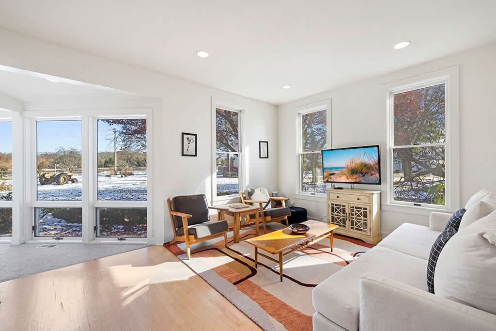 Spacious living room with light hardwood floors, white walls, and large windows offering bright outdoor views. A white sofa, two armchairs, and a coffee table are arranged on a geometric rug.