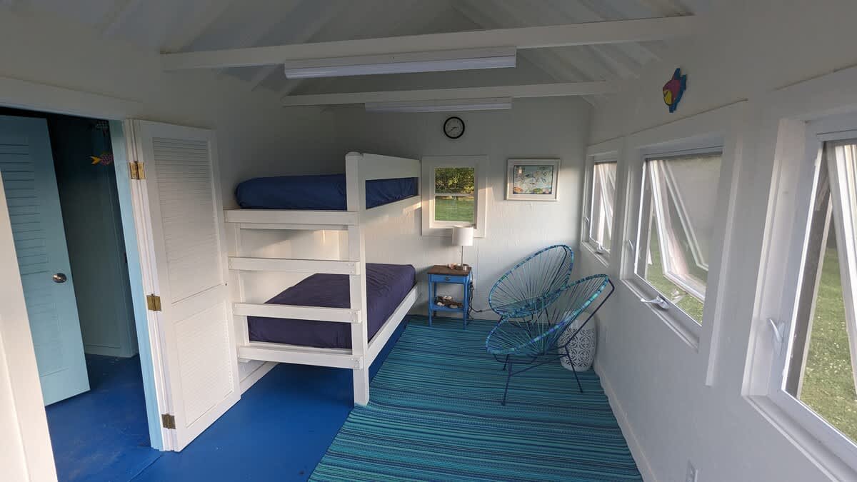 A bright and airy room with crisp white walls and a vibrant blue floor, featuring a sturdy white bunk bed with matching blue bedding. Two modern blue string chairs and multiple windows offering views of the outdoors complete the cheerful space.