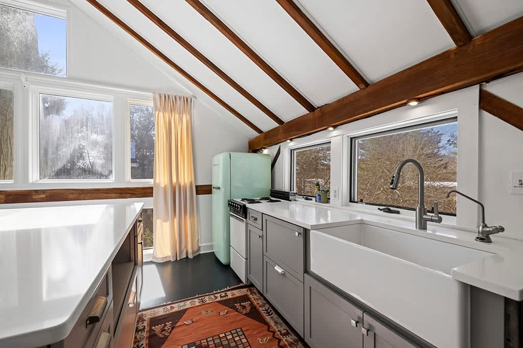 Modern kitchen with grey cabinetry, white countertops, and a large farmhouse sink, illuminated by natural light from multiple windows. A vintage-style mint green refrigerator and a stove are visible, along with a colorful patterned rug on the dark floor.